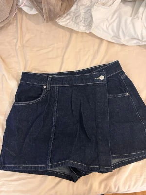 Mörkblå jeanskjol från Zara - Säljer en mörkblå jeanskjol från Zara i klassisk denim med vita kontrastsömmar. Kjolen har fickor fram och bak, bälteshällor och stängs med knapp och dragkedja. Perfekt för dig som gillar en enkel men snygg look.