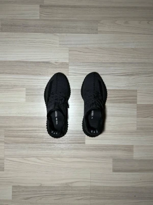 Svarta Adidas Yeezy Boost 350 V2 - Säljer ett par svarta Adidas Yeezy Boost 350 V2 med stickad ovandel och klassisk sula med boost-dämpning. Skorna har snörning, rund tå och diskreta ränder på sidan. Perfekta för dig som vill ha en clean och modern sneaker med streetwear-vibe. OBS: Köpta från Vinted! Ingen garanti 