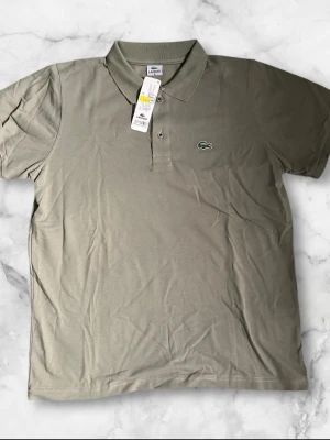 Olivgrön pikétröja från Lacoste - Säljer en klassisk olivgrön pikétröja från Lacoste med den ikoniska krokodilloggan på bröstet. Tröjan har krage, knappar framtill och korta ärmar. Perfekt för en clean och sportig look.