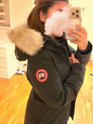 Svart Canada Goose dunjacka med päls - Säljer en svart Canada Goose dunjacka med stor fluffig päls runt huvan och klassisk logga på ärmen. Jackan har dragkedja och knappar framtill, samt flera praktiska fickor. Perfekt för kalla vinterdagar och riktigt snygg streetstil. Storlek XS men funkar även som S. Pris kan diskuteras.