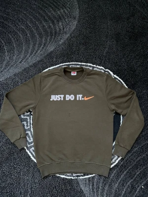 Olivgrön Nike sweatshirt Just Do It - Säljer en olivgrön sweatshirt från Nike i 100% bomull. Tröjan har trycket 'JUST DO IT.' och en orange swoosh på bröstet. Klassisk rund halsringning och ribbade muddar vid ärmar och nederkant. Perfekt för dig som gillar sportig och enkel stil.