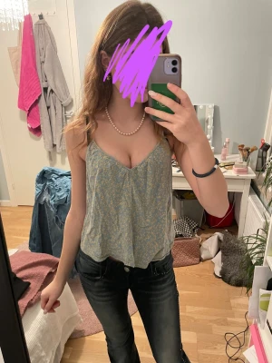 Blommig blå linnetopp - Söt blå linnetopp med små blommor i vitt och gult. Toppen har tunna axelband och en avslappnad, luftig passform som är perfekt för varma dagar. Materialet känns lätt och skönt mot huden, och toppen har en v-ringad urringning framtill.