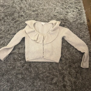 Beige ribbad kofta med volangkrage - Supersöt beige ribbad kofta med stora volangdetaljer runt kragen och knappar framtill. Långärmad och croppad modell som ger en trendig look. Perfekt att styla med jeans eller kjol för en chic vibe.