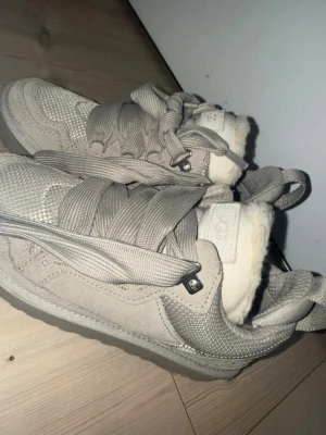 Gråbeiga sneakers från UGG med fårfoder - Säljer ett par gråbeiga sneakers från UGG i storlek 38. Skorna har breda snören, detaljer i mocka och mesh samt ett fluffigt fårfoder på insidan som gör dem extra mysiga. Perfekta för dig som vill ha både stil och komfort under kyligare dagar. Använda max 3 gånger. Nypris: 3800 från Vestiarie Collective. Färgens namn: Pumice