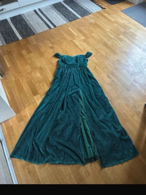 Mörkgrön glittrig balklänning - Supersnygg mörkgrön långklänning med glittereffekt och offshoulder-design. Klänningen har en markerad midja, slits på ena sidan och ett svepande fall. Perfekt för dig som vill sticka ut på bal eller fest! Materialet känns lyxigt och skimrande. Endast använd ca 1,5 timmar!