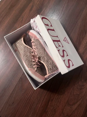 GUESS Sneakers - Sneakers från Guess i beige och ljusrosa med monogrammönster över hela skon. Skorna har ljusrosa snörning och är gjorda i ett canvas liknande tyg. Materialet är textil med gummi detaljer. Aldrig använda! Storlek 37. Säljs på flera sidor, först till kvarn. 