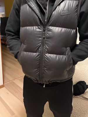 Svart dunväst från Nike - Säljer en svart, quiltad dunväst från Nike med diskret logga på bröstet. Västen har dragkedja framtill, två fickor och en hög krage. Perfekt för lager-på-lager och funkar till både streetwear och chill dagar. Materialet är syntet och dun för extra värme. Knappt använd nyskick nästan, modellen på bilden är 170cm