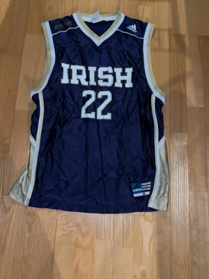 Adidas Notre Dame basketlinne blå/guld - Snyggt basketlinne från Adidas med Notre Dame IRISH och nummer 22 i vitt och guld på marinblå botten. Klassisk sportig look med breda axelband och kontrastpaneler i guld på sidorna. Perfekt för dig som gillar collegebasket och retrostil.