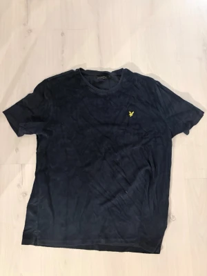 Mörkblå t-shirt från Lyle & Scott - Mörkblå t-shirt från Lyle & Scott med relaxed fit och klassisk rund hals. T-shirten har korta ärmar och en gul broderad logga på bröstet. Perfekt för en chill och stilren look.