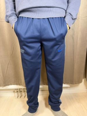 Nike Dri-Fit blå mjukisbyxor XL Barn - Blå Nike Dri-Fit mjukisbyxor med elastisk midja och vita samt blå ränder längs sidorna med små swoosh-loggor. Byxorna har normal passform och är gjorda i ett syntetmaterial som andas. Perfekta för träning eller chill, med fickor på sidorna och broderad Nike-logga.