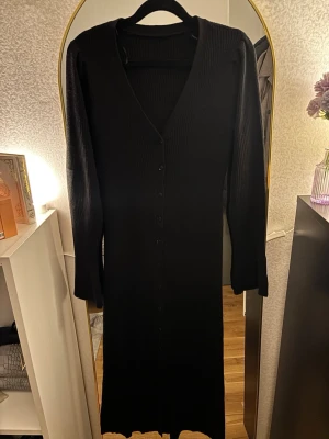 Svart ribbad långklänning med knappar - Säljer en svart långklänning i ribbat material med v-ringning och långa ärmar. Klänningen har knappar hela vägen framtill och är figurnära i modellen. Perfekt för dig som gillar stilrena och enkla plagg med en twist.