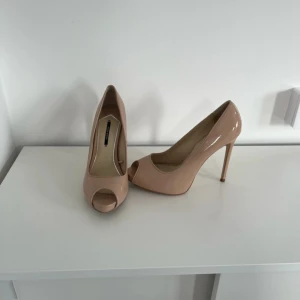Beige pumps med klack från Zara  - Snygga beige pumps med hög klack och öppen tå. Tillverkade i ett glansigt syntetmaterial som ger en elegant look. Perfekta för att lyfta din outfit med stil.
