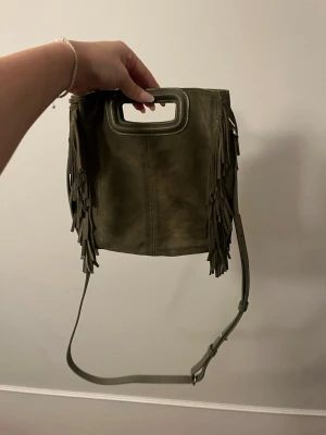 Make väska i grön - Säljer en beige axelväska i mocka från Maje med snygga fransar på sidorna och ett stilrent handtag upptill. Väskan har en avtagbar axelrem och en cool, fyrkantig form. Perfekt för dig som vill ha en unik accessoar med lite extra edge.