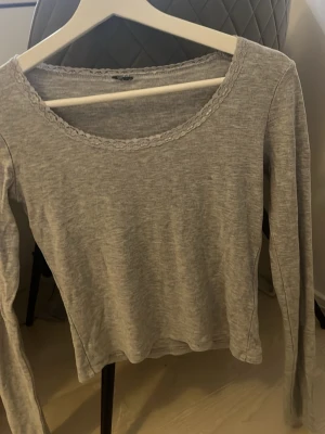 Grå långärmad topp med spetskant - Säljer en enkel och stilren grå långärmad topp med rund halsringning och fin spetskant runt halsen. Toppen är i mjukt material och har en normal passform som funkar till många olika looks.