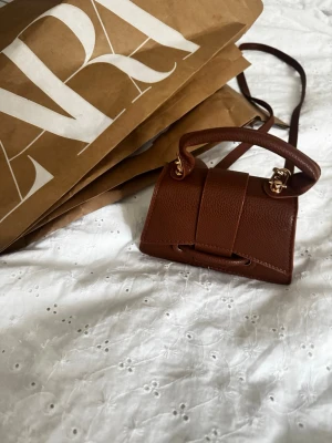 Brun minihandväska från Zara - Säljer en liten brun handväska från Zara i stilren design. Väskan har ett justerbart axelband, guldfärgade metalldetaljer och ett topphandtag. Tillverkad i strukturerat syntetmaterial som liknar skinn. INTE ANVÄND 