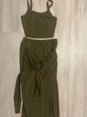 Olivgrön ribbad kjol med knyt - Säljer en olivgrön kjol i ribbat material med smala axelband och croppad passform. Kjolen har ett snyggt knyt framtill som ger en extra detalj. Perfekt att styla med ett par fina skor för en trendig look.