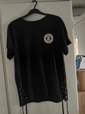 Svart t-shirt med snörning och patch - Svart t-shirt med cool snörning i sidorna och rund hals. På bröstet finns en beige patch med texten 'Colorful Life 1984' och på ena ärmen sitter en röd broderad detalj. T-shirten har korta ärmar och är gjord i mjuk bomull.