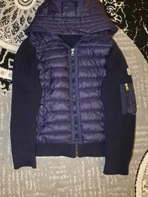Mörkblå Moncler pufferjacka med huva - Säljer en mörkblå pufferjacka från Moncler med stickade ärmar och vadderad kropp. Jackan har huva, dragkedja framtill och en ficka med dragkedja på ärmen. Klassisk Moncler-logga på ärmen och coolt serietryck på insidan. släng in din bud. Den passar på båda till killar och tjejer 