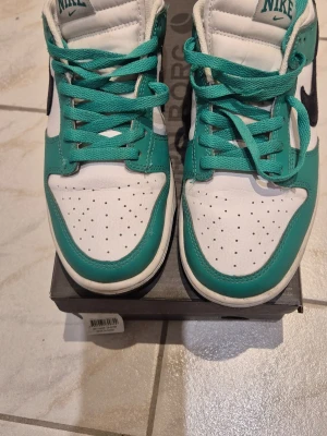 Nike Dunk Low vit/turkos - Säljer ett par Nike Dunk Low sneakers i vitt och turkost med svart swoosh. Skorna har klassisk snörning, perforerad tåbox och lågt skaft. Ovandelen är i skinn och innerfodret är i mesh för extra komfort. Perfekta för dig som gillar streetwear och vill sticka ut.