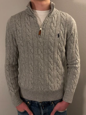 Grå stickad tröja Polo Ralph Lauren - Säljer en grå kabelstickad Ralph Lauren tröja med half zip och hög krage. Tröjan har lång ärm, ribbade muddar och klassisk broderad logga på bröstet. Modellen på bilden är 183cm så den passar bäst för personer som är 175 till 180cm