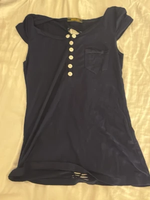 Mörkblå topp med knappar från MNG Suit - Supersnygg mörkblå topp från MNG Suit med vita knappar framtill, liten bröstficka och coolt utskuret mönster längs ryggen. T-shirten är ärmlös och har en tight passform som sitter snyggt på kroppen. Perfekt för dig som gillar unika detaljer!