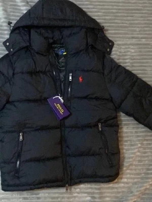 Svart pufferjacka från Polo Ralph Lauren - Säljer en svart pufferjacka från Polo Ralph Lauren med huva och dragkedjor på fickorna. Helt ny aldrig använt som ni kan se på bilden säljer jätte billigt pga behöver pengar till studenten så passa på nu när de är vinter skriv om ni har funderingar 