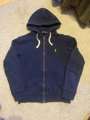 Polo Ralph Lauren zip hoodie – M - Mörkblå zip hoodie från Polo Ralph Lauren. Klassisk modell med huva, fickor framtill och broderad logga på bröstet.  ▪️ Storlek: M ▪️ Skick: Bra ▪️ Notering: Litet hål vid armvecket (se bild) ▪️ Färg: Mörkblå  Hel och fräsch i övrigt. Redo att användas 🌟 Skicka gärna meddelande vid frågor!