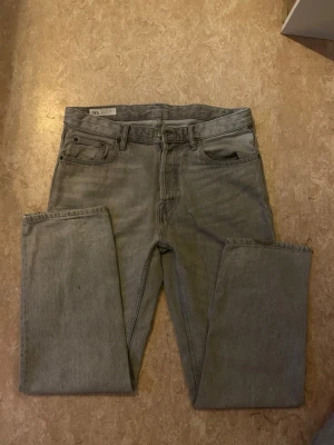 Grå raka jeans från Zara, strl 42 - Grå jeans från Zara i rak modell med klassisk femficksdesign och bälteshällor. Jeansen är tillverkade i bomull och har en enkel, stilren look utan slitningar eller dekorationer. Passar dig som gillar en clean och avslappnad vibe. Använd bara några gånger och är i väldigt bra skick utan defekter. Kom med bud!