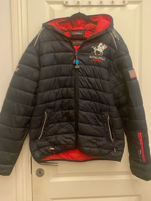 Svart pufferjacka Royal Polo - Snygg svart pufferjacka från Royal Polo i storlek L med broderad logga och amerikansk flagga på ärmen. Jackan har röd insida, dragkedjor på fickorna och detaljer i vitt och rött. Perfekt för kalla dagar med sin vadderade design och sportiga look.