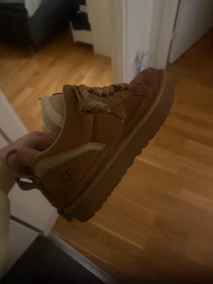 Bruna sneakers från UGG med foder - Säljer ett par bruna sneakers från UGG med grov sula och breda snören. Skorna har mysigt vitt foder på insidan och är gjorda i mocka och textil. Perfekta för kyligare dagar och har en snygg, chunky look.