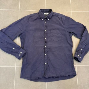 Blå skjorta från J.Lindeberg - Säljer en stilren blå skjorta från J.Lindeberg i slim fit-modell. Skjortan har klassisk krage med knappar, bröstficka och vita knappar längs hela framsidan. Tillverkad i ett mjukt bomullstyg och har långa ärmar.