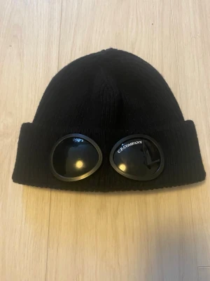 Svart mössa med goggles från C.P. Company - Säljer en svart ribbstickad mössa från C.P. Company med ikoniska goggles framtill. Mössan är i mjukt material och har en bred uppvikt kant. Perfekt accessoar för dig som vill sticka ut lite extra.
