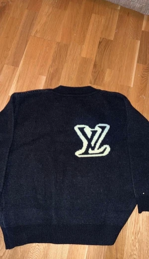  Louis Vuitton tröja - Säljer en svart stickad tröja från Louis Vuitton i storlek M. Tröjan har ett stort vitt LV-logo broderat på bröstet och ribbade muddar vid ärmslut och nederkant. Klassisk rund halsringning och loose fit för en chill vibe.