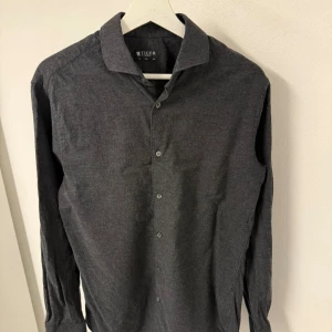 Mörkgrå skjorta Tiger of Sweden - Snygg mörkgrå skjorta från Tiger of Sweden i slim fit. Skjortan har klassisk krage, långa ärmar och knappar framtill. Materialet är mjukt och känns som bomull, perfekt för dig som gillar stilrena och enkla plagg med en modern passform. Storlek 40