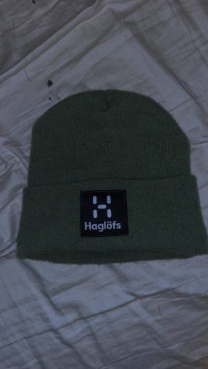 Grön stickad mössa från Haglöfs - Snygg grön stickad mössa från Haglöfs med uppvikt kant och svart patch med vit logga framtill. Perfekt för kalla dagar och enkel att matcha med olika outfits. Klassisk och stilren design som funkar året runt.