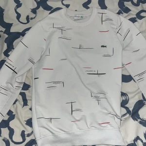 Vit sweatshirt från Lacoste med print - Vit sweatshirt från Lacoste med abstrakt print i svart och rött. Tröjan har långärmad design, rund hals och den klassiska gröna krokodilloggan på bröstet. Perfekt för dig som gillar stilrena och sportiga plagg med en twist.