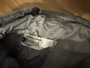 Grå pufferjacka från C.P. Company - Säljer en grå pufferjacka från C.P. Company med huva och coolt glaslins-detalj på ärmen. Jackan har quiltad design, dragkedja framtill och två fickor med dragkedja. Perfekt för dig som vill ha en stilren och varm jacka med streetkänsla.