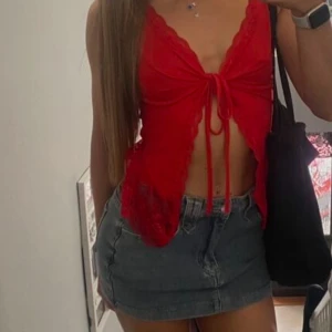 Röd omlottopp med spets och knyt - Säljer en röd omlottopp med spetsdetaljer längs kanterna och knyt framtill. Toppen är croppad och har ärmlös design med v-ringad hals. Perfekt för dig som gillar att sticka ut med färg och snygga detaljer.