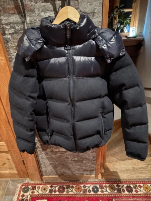 Mörkblå pufferjacka från Polo Ralph Lauren - Mörkblå pufferjacka från Polo Ralph Lauren med både matt och glansig finish. Jackan har hög krage, avtagbar huva och två sidofickor med dragkedja. Materialmix av ull och polyester, quiltad design och hel dragkedja framtill. Perfekt för kalla dagar.