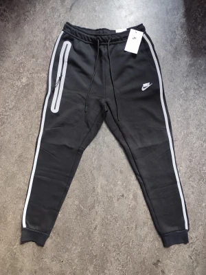 Svarta Nike mjukisbyxor med reflex - Svarta mjukisbyxor från Nike med vita reflexdetaljer längs benen och runt dragkedjan på fickan. Byxorna har snörning i midjan, smal passform och ribbade muddar vid bensluten. Perfekt för dig som gillar sportig stil och vill synas i mörkret.