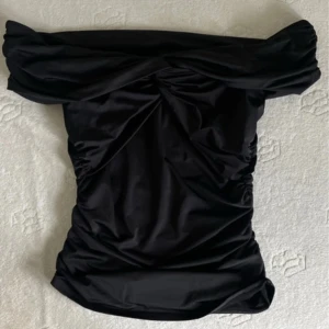 Svart offshoulder tröja Gina Tricot - Supersnygg svart tröja från Gina Tricot i offshoulder-modell. Den har draperade detaljer och en figurnära passform som framhäver siluetten. Materialet är mjukt och har en lätt glansig finish, perfekt för dig som gillar trendiga och stilrena looks.