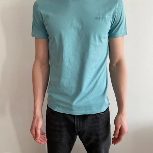 Ljusblå t-shirt från BOSS - Säljer en stilren ljusblå t-shirt från BOSS i mycket bra skick. Den är kortärmad och har en normal passform, perfekt för både vardag och fest. Materialet känns mjukt och skönt mot huden. Diskret BOSS-logga på bröstet ger en extra touch av stil. Passar perfekt till jeans eller shorts!