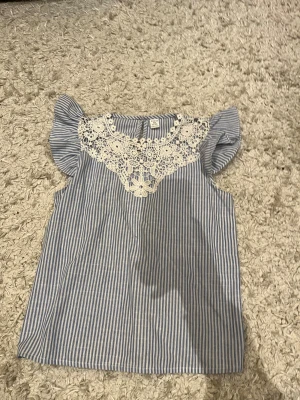 Randig blus med spets från SHEIN - Säljer en söt blus från SHEIN i storlek 11-12 år. Blusen är blå- och vitrandig med volangärmar och har en vit spetsdetalj med blommönster vid halsen. Perfekt för dig som gillar romantiska detaljer och fräscha färger.