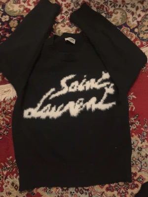 Svart stickad Saint Laurent tröja - Svart stickad tröja från Saint Laurent med vit, fuzzy logotyp på bröstet. Tröjan har rund hals och långa ärmar, perfekt för dig som gillar exklusiva märken och vill ha en cool statement piece i garderoben. Pris kan såklart diskuteras 