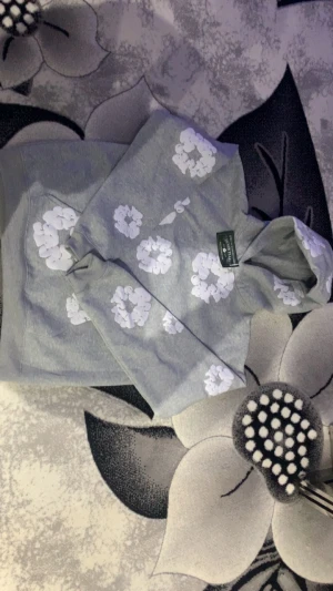 Grå hoodie med vita blommor Denim Tears - Säljer en grå hoodie från Denim Tears i storlek L, prydd med vita blommor över hela tröjan. Hoodien har huva och en klassisk känguruficka framtill. Perfekt för dig som gillar streetwear med unik design och vill sticka ut lite extra.