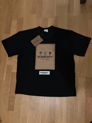 Svart Burberry t-shirt med beige print - Snygg svart t-shirt från Burberry med ett stort beige tryck på bröstet och vit patch med Burberry-logga nedanför. Klassisk rund hals och korta ärmar. Materialet är mjuk bomull och passformen är normal. Perfekt för dig som gillar exklusiva streetwear-vibbar.