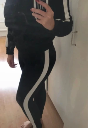 SET! Leggings med hoodie från H&M - Svart hoodie med vita ränder längs ärmarna och huva. Tröjan har snörning runt halsen och en stor magficka framtill samt med tillhörande svarta leggings med en bred vit rand längs utsidan. Sitter tight och har en sportig vibe, perfekta för en avslappnad och sportig stil