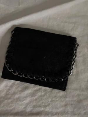 Svart mocka plånbok med kedjedetalj - Snygg svart plånbok i mocka med silverfärgad kedja runt kanten. Plånboken har flera fack för kort och stängs med magnetknapp. Perfekt storlek för att ha med sig överallt och ger en cool vibe med kedjedetaljen.