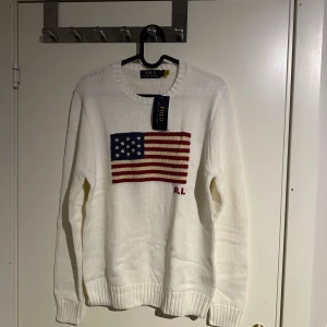 Vit stickad tröja Polo Ralph Lauren - Vit stickad tröja från Polo Ralph Lauren med amerikansk flagga i blått, rött och vitt på bröstet samt RL-broderi. Rund hals och ribbade muddar vid ärmslut och nederkant. Perfekt för dig som gillar klassisk och clean stil.