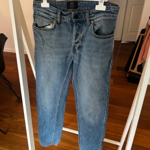 Blå jeans från Neuw Stockholm - Säljer ett par klassiska blå jeans från Neuw Stockholm. Modellen har fem fickor, raka ben och normal passform. Jeansen är tillverkade i slitstarkt denim med snygga kontrastsömmar och knappstängning fram. Perfekt för dig som gillar enkel och tidlös stil. Lite sliten vid hälen 
